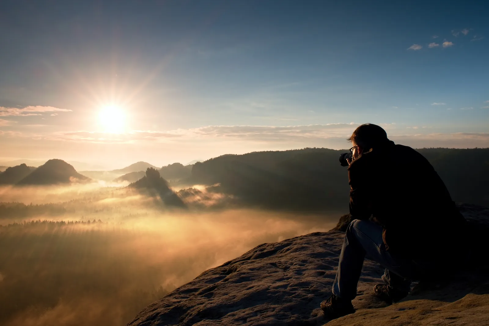 Fotógrafo sentado em uma rocha observando o nascer do sol sobre um vale coberto de névoa